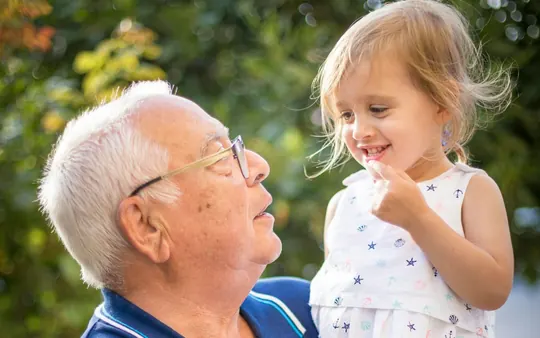Een opa in een blauwe polo houdt lachend zijn kleindochter vast